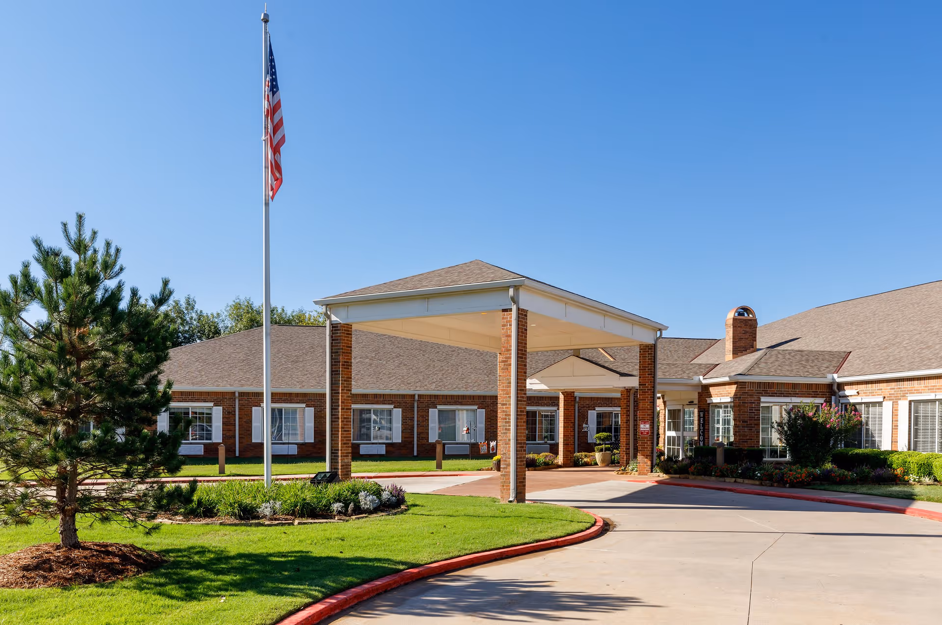 Exterior view of Rivermont Assisted Living and Memory Care facility showing a brick building with a covered entrance, an American flag on a flagpole, well-maintained green lawn, and clear blue sky.