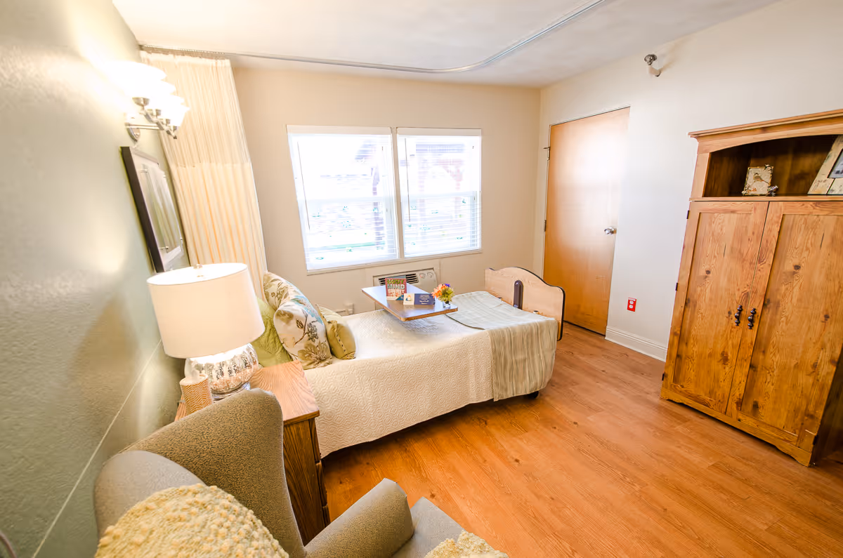 A cozy bedroom in a senior living facility featuring a single bed with a beige quilt and decorative pillows, a wooden nightstand with a lamp, a comfortable armchair with a cushion, a wooden wardrobe, and two windows with blinds allowing natural light to fill the room.