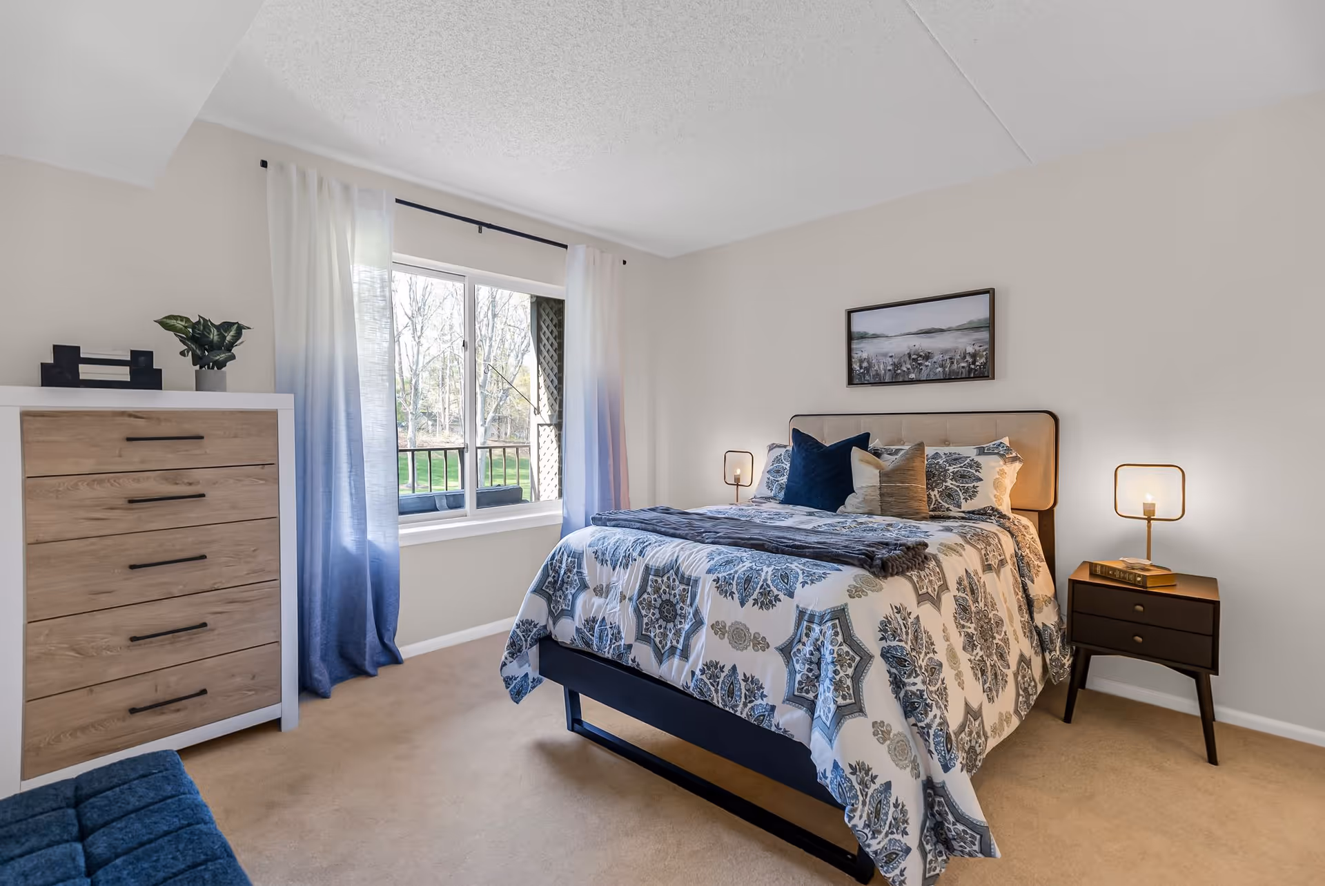 A bright bedroom with a large window covered by white and blue ombre curtains. The room features a bed with a patterned blue and white comforter and multiple pillows, a wooden dresser with black handles, a small dark nightstand with a modern lamp and a book, beige carpet, and a framed landscape painting on the wall above the bed.