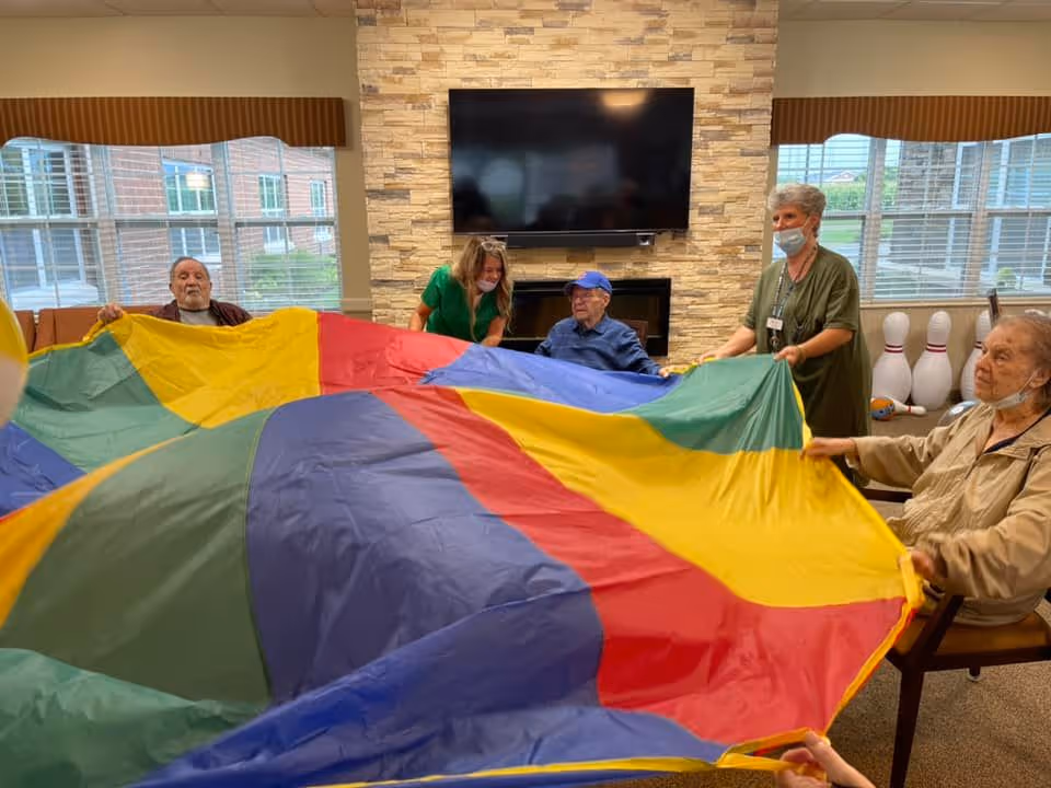 A group of elderly people and a caregiver holding the edges of a large colorful parachute in a cozy room with a stone fireplace and a large TV mounted above it. The room has large windows with brown valances and bowling pins in the background.