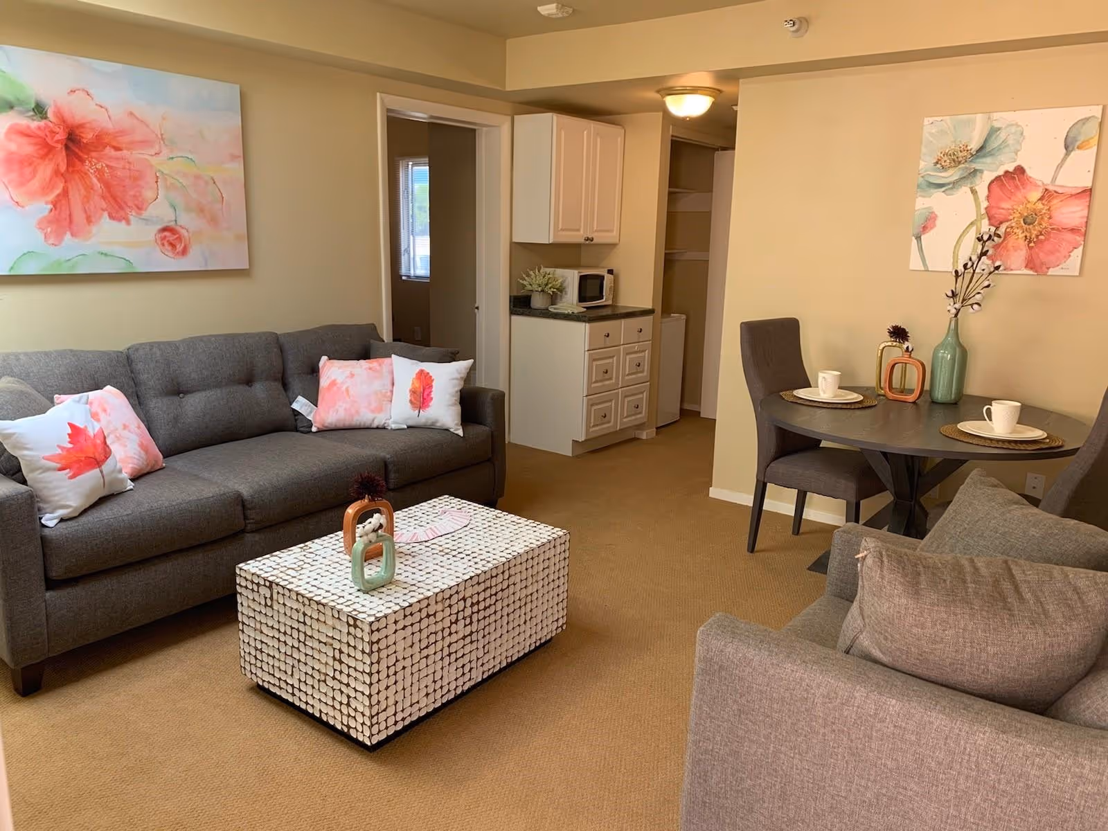 A cozy living room area with a gray sofa adorned with floral-themed pillows, a matching armchair, and a white patterned coffee table. There is a small round dining table with two chairs set with plates and cups. The walls are decorated with large floral paintings, and there is a small kitchenette area with cabinets, a microwave, and a small refrigerator visible in the background.