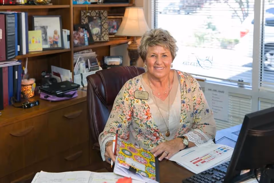 A smiling woman with short blonde hair sits at a desk in an office. She is wearing a floral blouse and a name tag that reads 'Las Fuentes Resort Village.' The desk has papers, a colorful folder, and a computer monitor. Behind her are shelves with books, framed photos, and a lamp. A window with blinds is visible in the background.