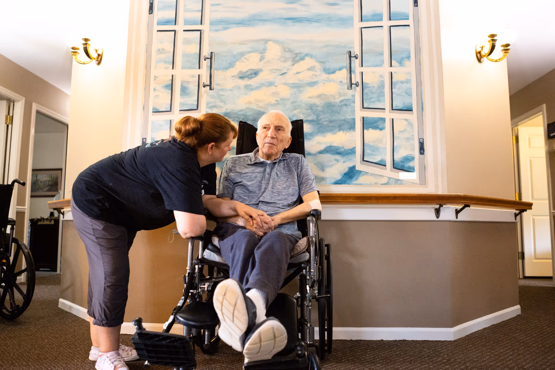 A caregiver is leaning down and holding hands with an elderly man seated in a wheelchair inside a senior living facility. Behind them is a wall mural depicting an open window with a view of a blue sky and clouds. The setting appears warm and supportive.