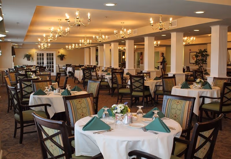 Spacious dining room with round tables set with white tablecloths, green folded napkins, floral centerpieces, chandeliers, and wooden chairs.