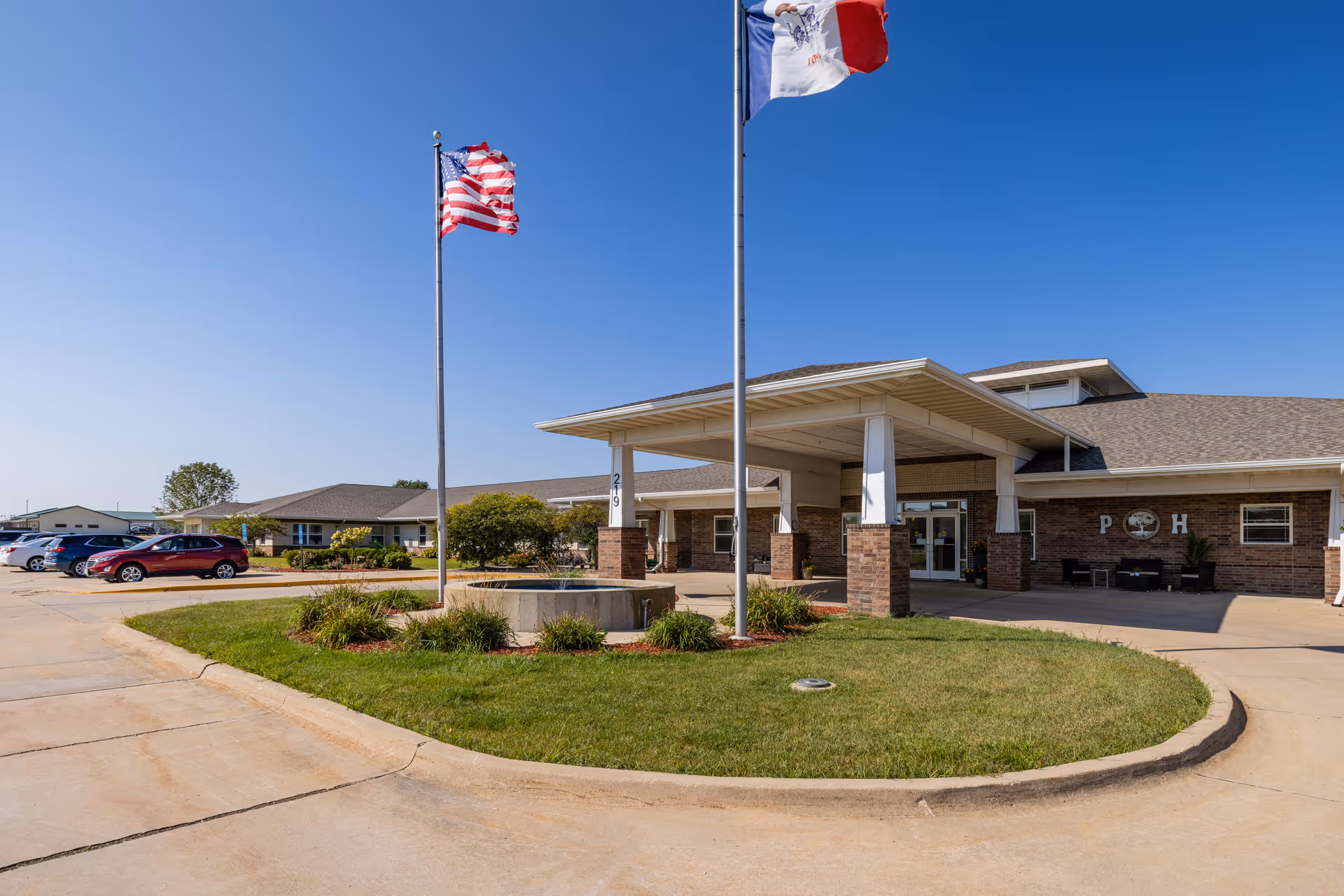 Front entrance of a single-story senior living facility with flagpoles, a circular driveway, and parked cars under a clear blue sky.