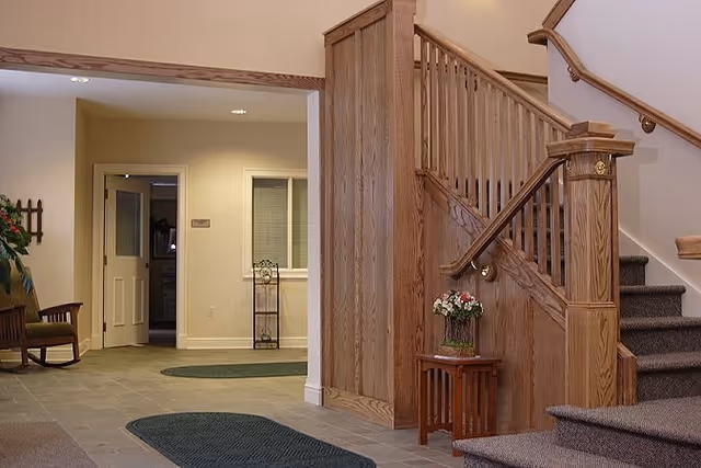 Interior view of a senior living facility showing a wooden staircase with carpeted steps, a small wooden table with a vase of flowers, a rocking chair, and a hallway leading to a door and window. The floor is tiled with green rugs placed on it.