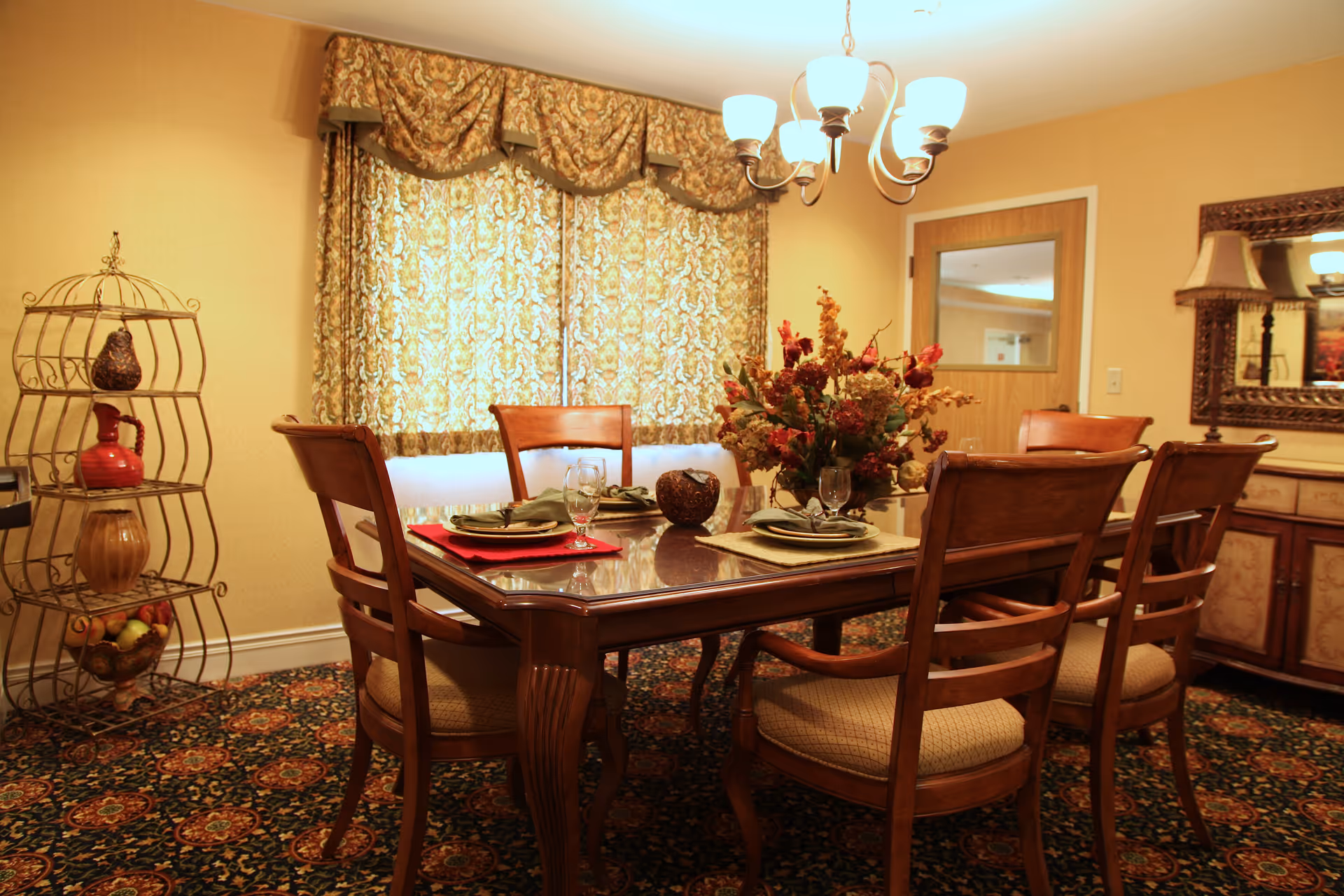 A warmly lit dining room with a wooden dining table set for four, featuring plates, napkins, and glasses. The room has patterned curtains covering a window, a decorative floral centerpiece on the table, a chandelier overhead, a sideboard with a large mirror, and a metal rack holding decorative items and fruit in the corner.