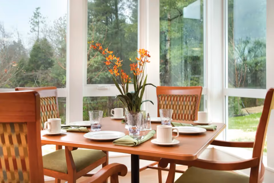 A dining table set for four with white plates, cups, glasses, and green napkins. A vase with orange flowers is in the center of the table. The room has large windows showing a green outdoor view.