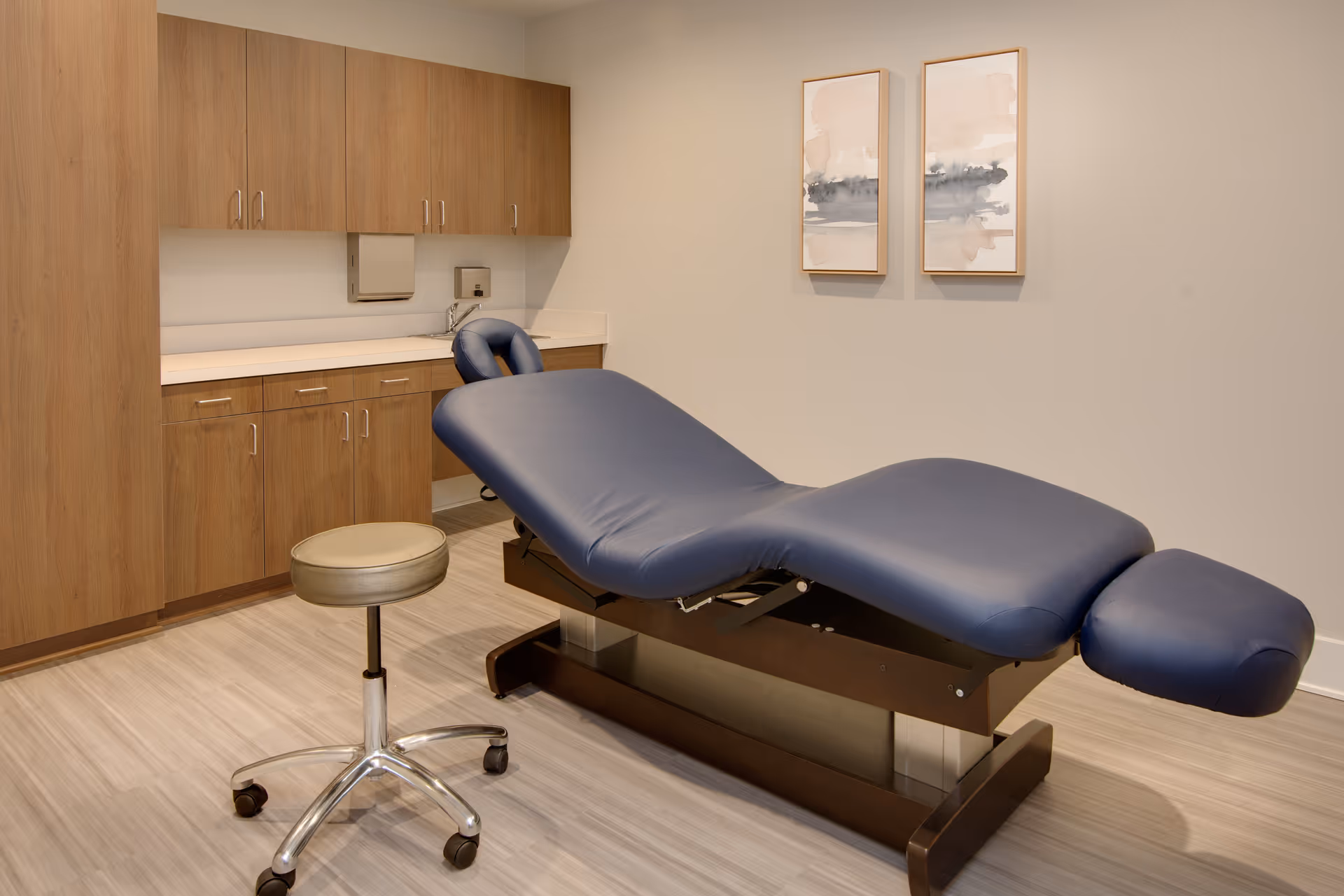 A treatment room with a blue adjustable medical examination table, a beige rolling stool, wooden cabinets along the wall, a countertop with a sink, and two abstract paintings hanging on a light-colored wall.