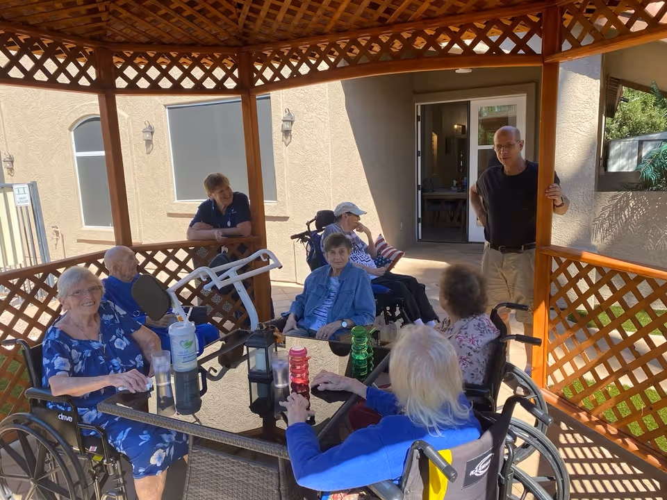 A group of elderly people, some in wheelchairs, sitting around a glass table under a wooden gazebo outdoors. Two caregivers are standing nearby, engaging with the group. The setting appears to be a courtyard or patio area of a senior living facility.