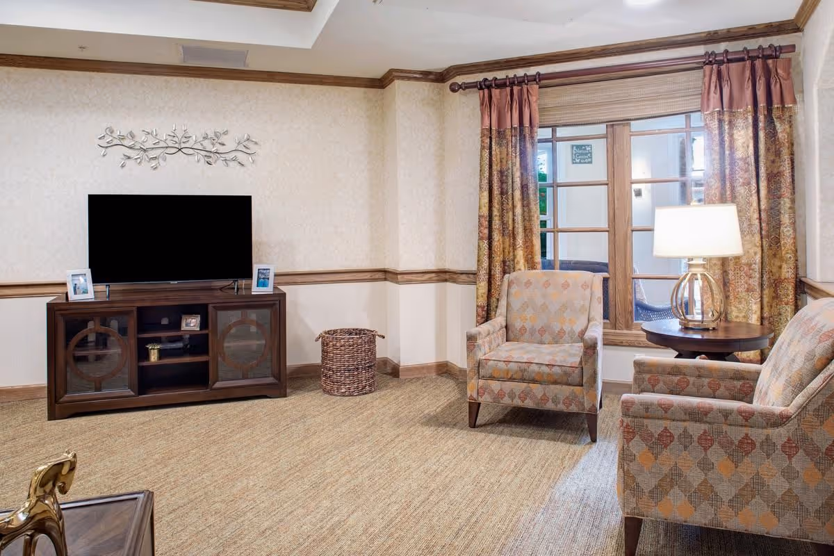 A cozy living room area with two patterned armchairs facing a wooden TV stand with a flat-screen television. The room features beige walls with wooden trim, a decorative metal wall art above the TV, a woven basket on the floor, a round wooden side table with a lamp, and a window with patterned curtains.