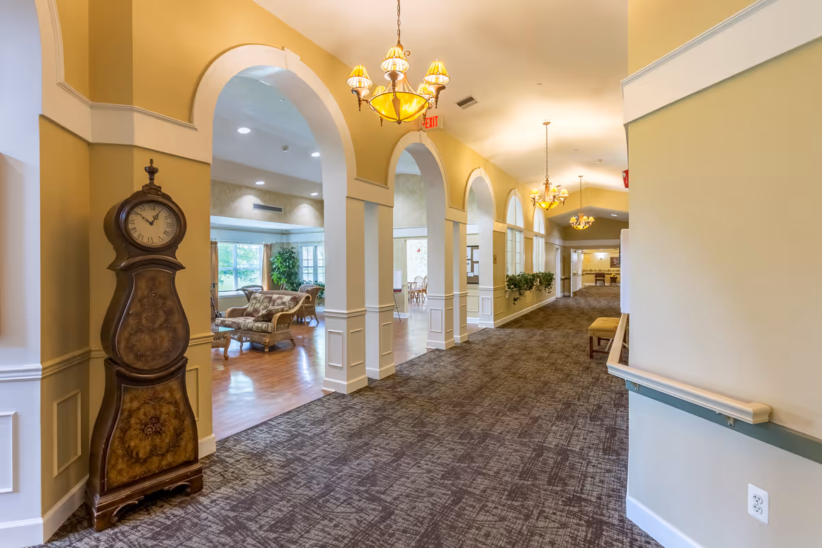 A well-lit hallway in a senior living facility with arched openings leading to a sitting area furnished with wicker chairs and a sofa. The hallway has patterned carpet, yellow walls with white trim, and multiple chandeliers hanging from the ceiling. A tall antique-style clock is positioned near one of the arches.