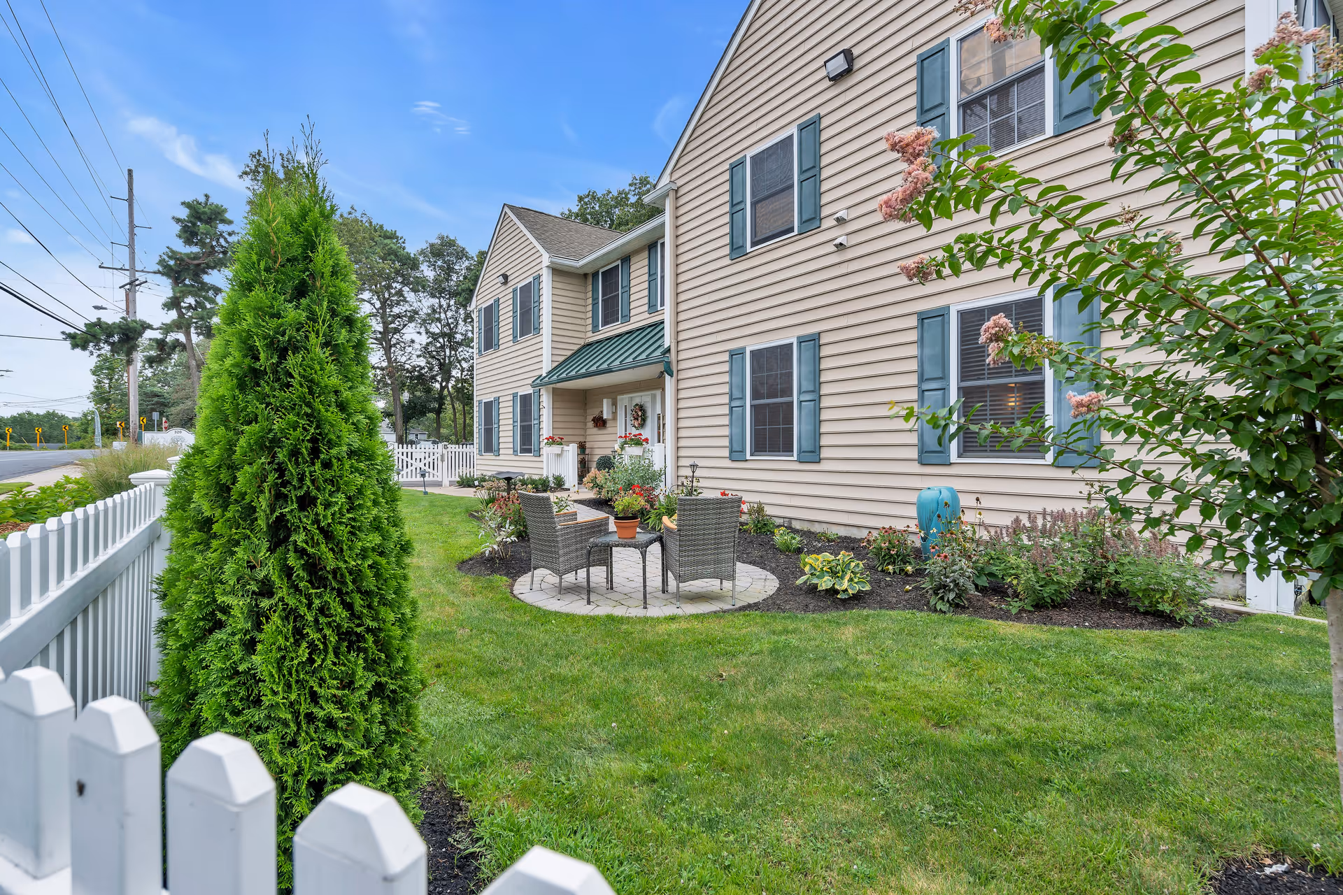 Outdoor view of a senior living facility named Serenity Gardens at Brick, showing a beige two-story building with green shutters and a small patio area with two chairs and a table surrounded by a well-maintained garden and white picket fence.