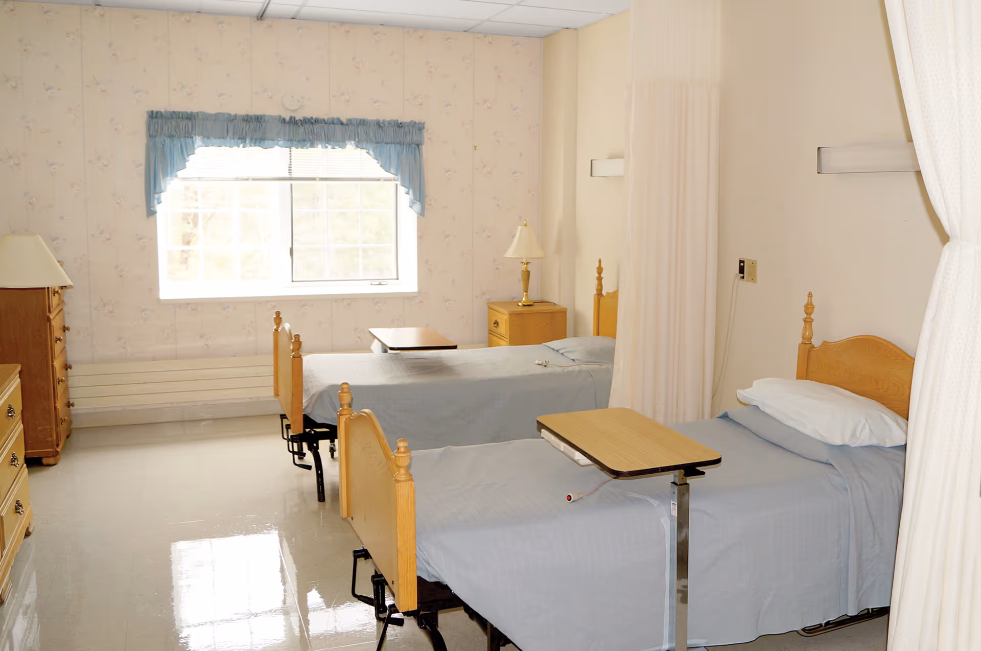 A room with two hospital-style beds separated by a curtain. Each bed has a light blue blanket and a pillow. There are two wooden nightstands with lamps, a window with blue valance curtains, and a wooden dresser. The floor is shiny and reflective.