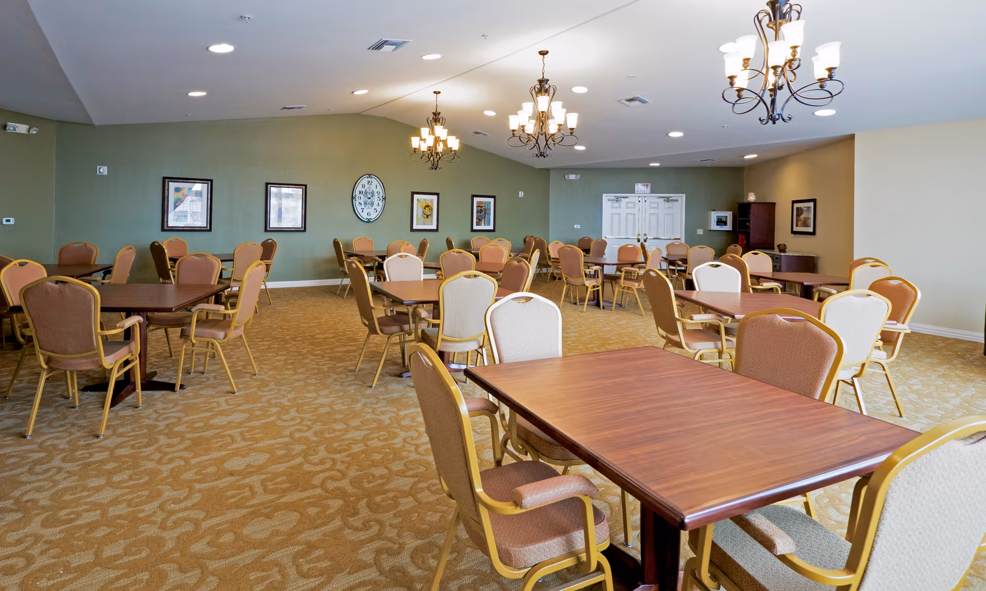 A spacious dining room with multiple square wooden tables and cushioned chairs arranged neatly. The room has beige patterned carpet, green and beige walls adorned with framed pictures and a large clock. Three chandeliers hang from the ceiling providing warm lighting.