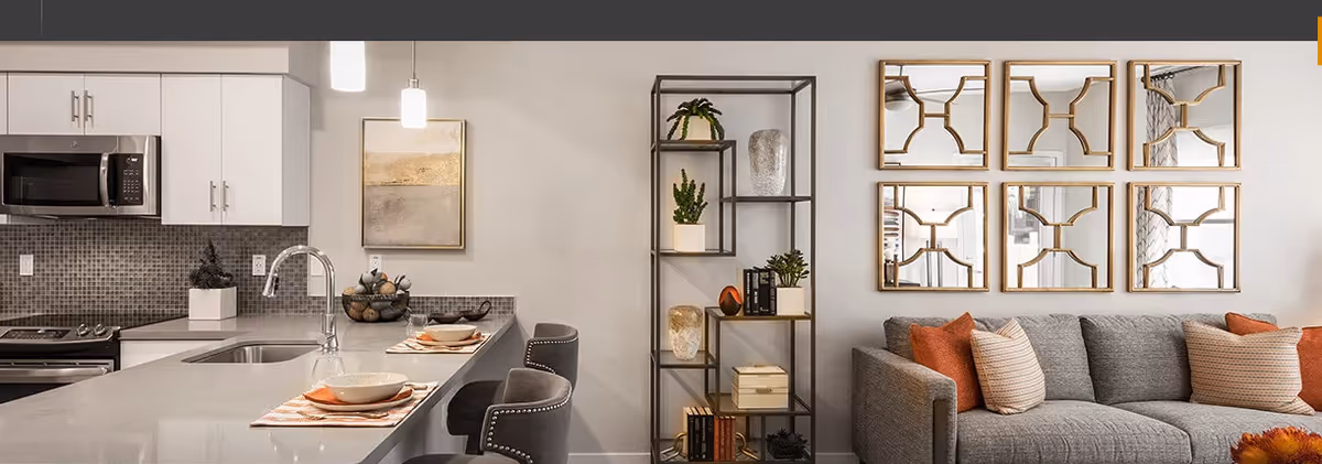 Open-plan living area with a modern kitchen island and bar stools beside a grey sofa, shelving, and decorative wall mirrors.