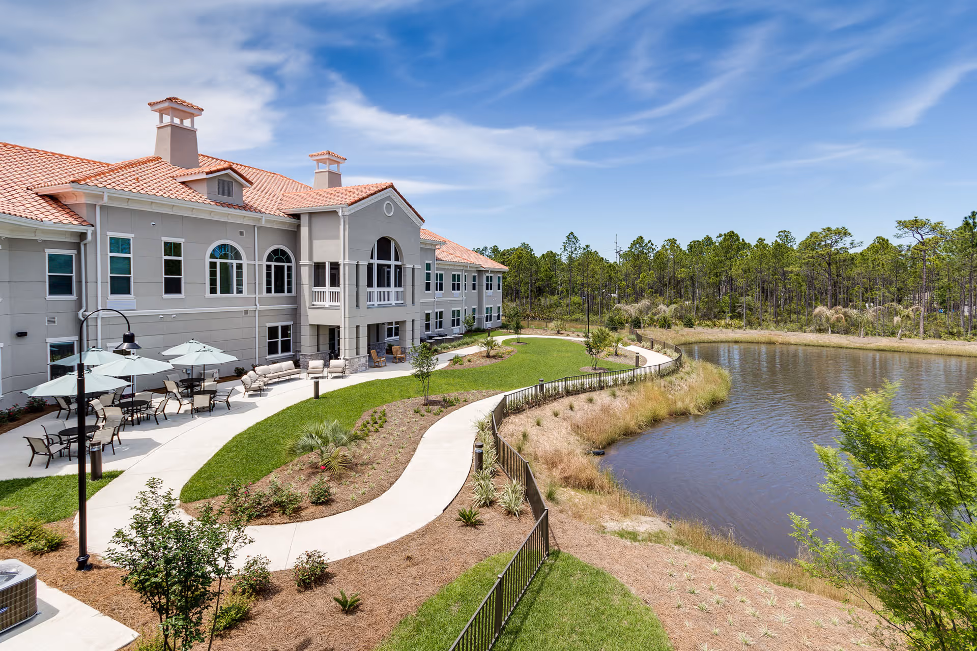 Two-story beige building with a red tile roof, outdoor seating, winding walkway and a pond under a blue sky.