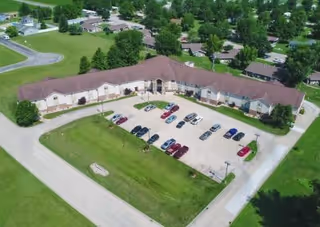 Aerial view of a single-story senior living facility named Heritage Woods of Flora, showing a large U-shaped building with a brown roof, surrounded by green lawns and trees. There is a parking lot in front of the building with several cars parked. Residential houses and streets are visible in the background.