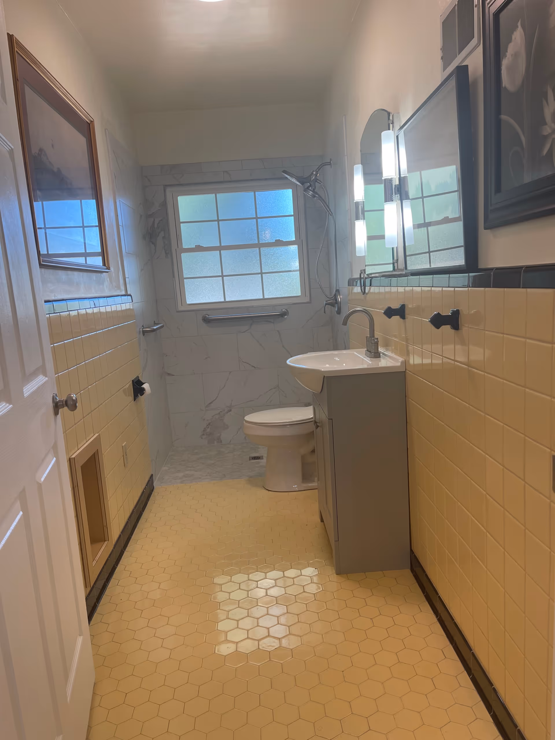 A narrow bathroom with yellow hex tile floor and yellow tiled walls, featuring a sink vanity, toilet, and a shower beneath a frosted window.