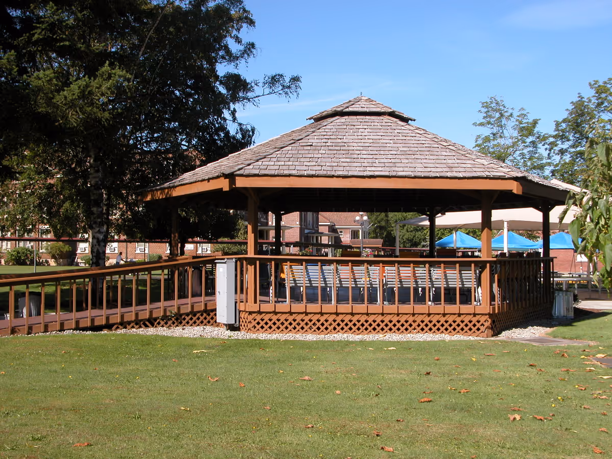 A wooden gazebo with a shingled roof situated on a grassy lawn with trees and buildings in the background under a clear blue sky.