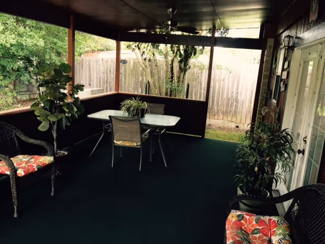 Covered outdoor patio area with a green carpet, a glass-top table with three chairs, two wicker chairs with floral cushions, several potted plants, and a wooden fence in the background.