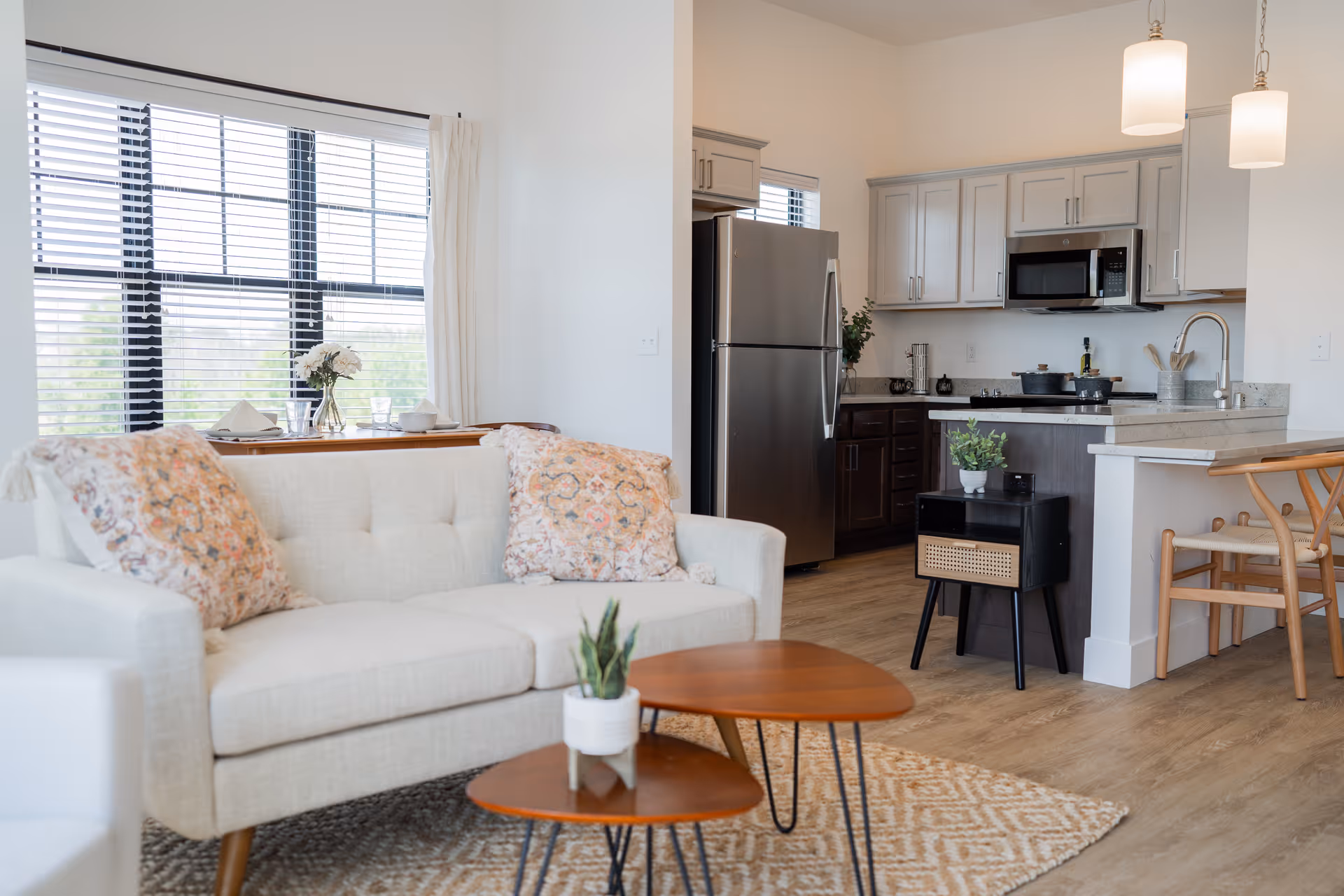 Bright open-concept living room and kitchen with a light sofa, patterned pillows, wooden coffee tables, and stainless-steel refrigerator.
