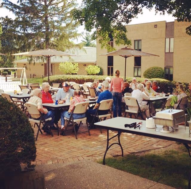 A group of elderly residents seated at outdoor tables under umbrellas on a patio beside a brick building.