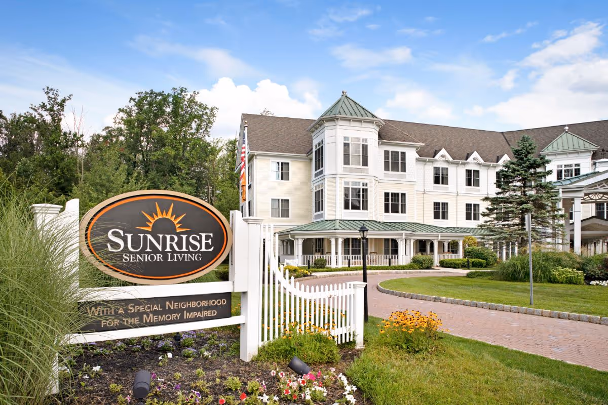 Exterior view of Sunrise Senior Living facility with a large sign in the foreground that reads 'Sunrise Senior Living - With a Special Neighborhood for the Memory Impaired'. The building is a multi-story structure with white siding, green roofs, and multiple windows. There is a curved brick driveway, well-maintained landscaping, flowers, and trees under a partly cloudy blue sky.
