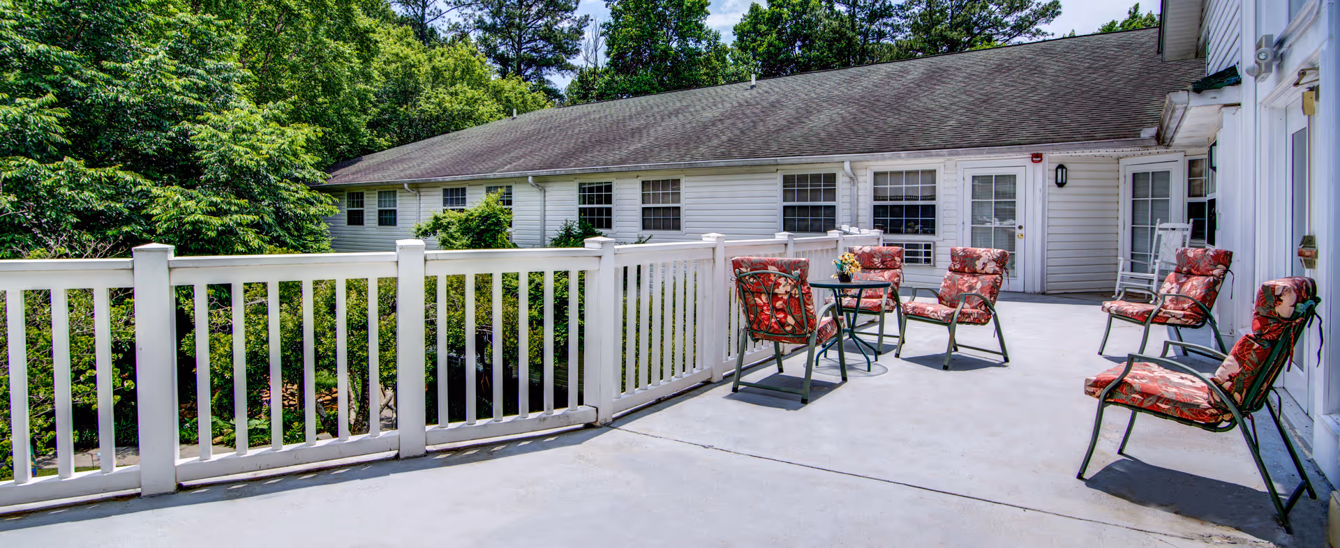 Sunny outdoor patio with white railing, cushioned chairs and small tables outside a single-story building.