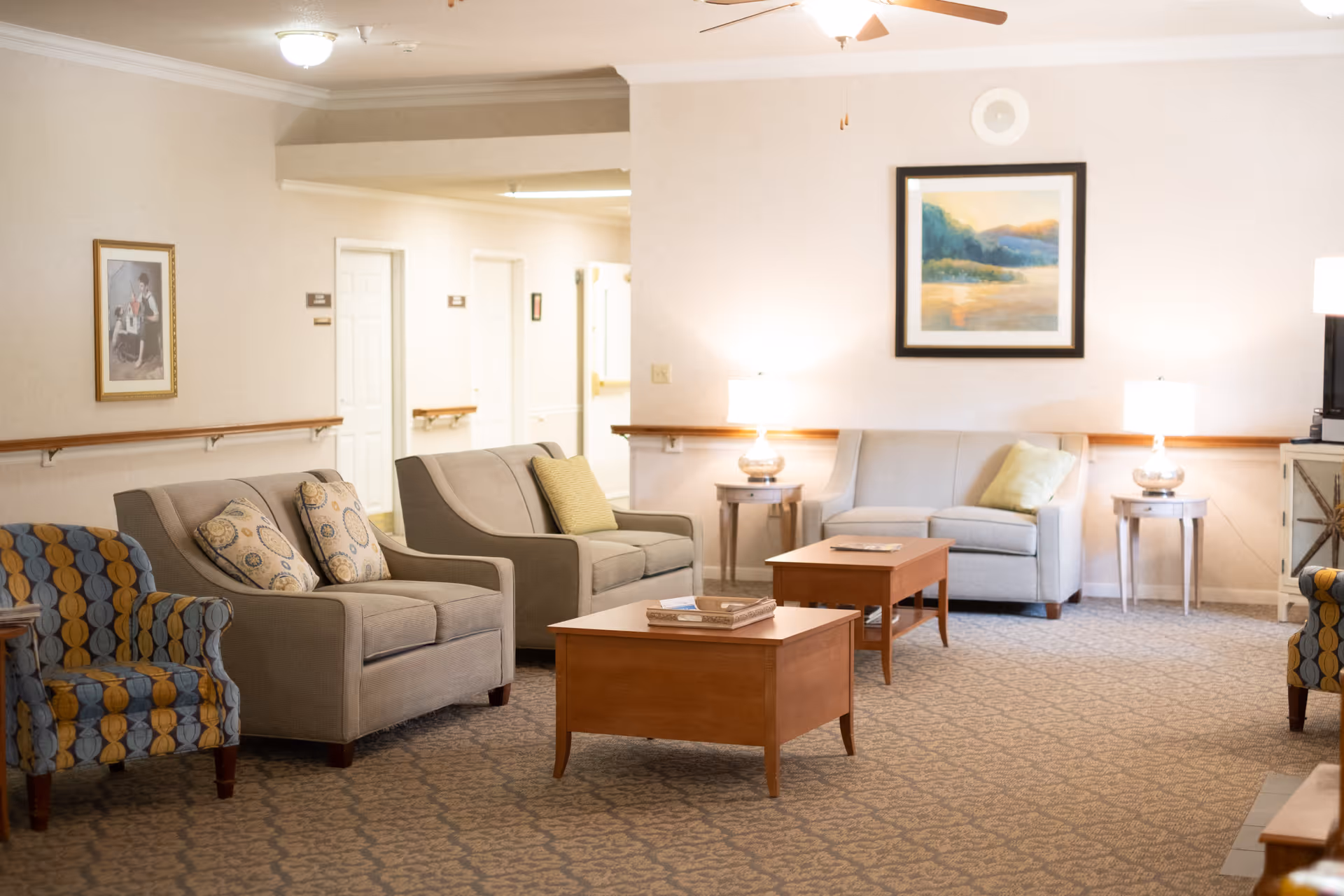 A well-lit assisted living facility common area with beige walls and carpeted floor. The room features several cushioned chairs and sofas with patterned and solid-colored pillows, two wooden coffee tables with books on them, two side tables with lamps, a ceiling fan, and framed artwork on the walls. A hallway with white doors is visible in the background.