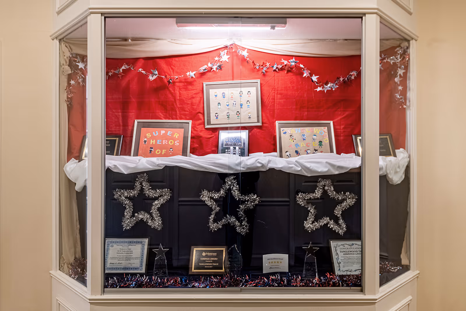 A display case decorated with red and white fabric and silver star garlands, featuring framed artwork with cartoon characters and text, along with several awards and certificates at the bottom.