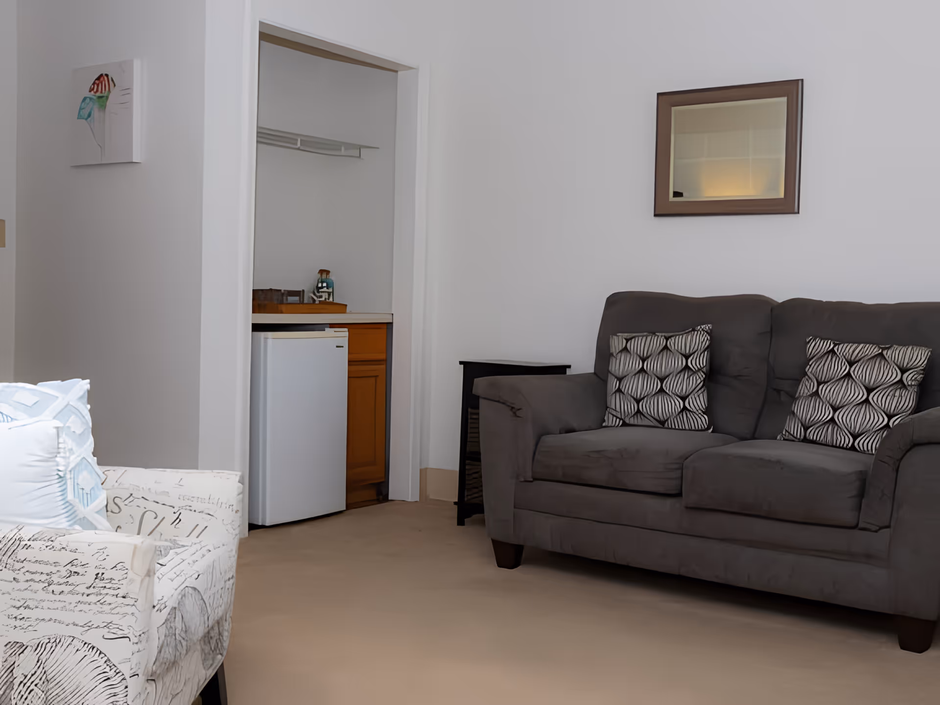 A cozy living room area with a dark gray couch adorned with two patterned pillows, a small black side table, and a white armchair with script and house patterns. A small white mini-fridge and wooden cabinet are visible through an open doorway. A framed mirror hangs on the wall above the couch, and a small piece of artwork is mounted on the adjacent wall.