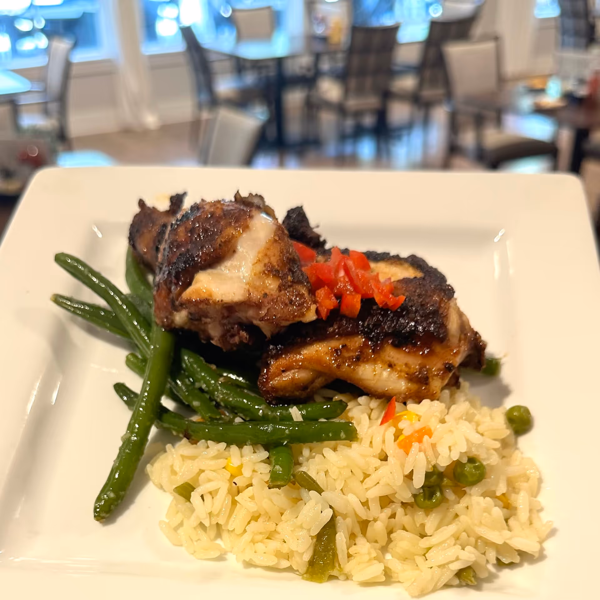 Grilled chicken with green beans and seasoned rice served on a square white plate in a dining room setting.