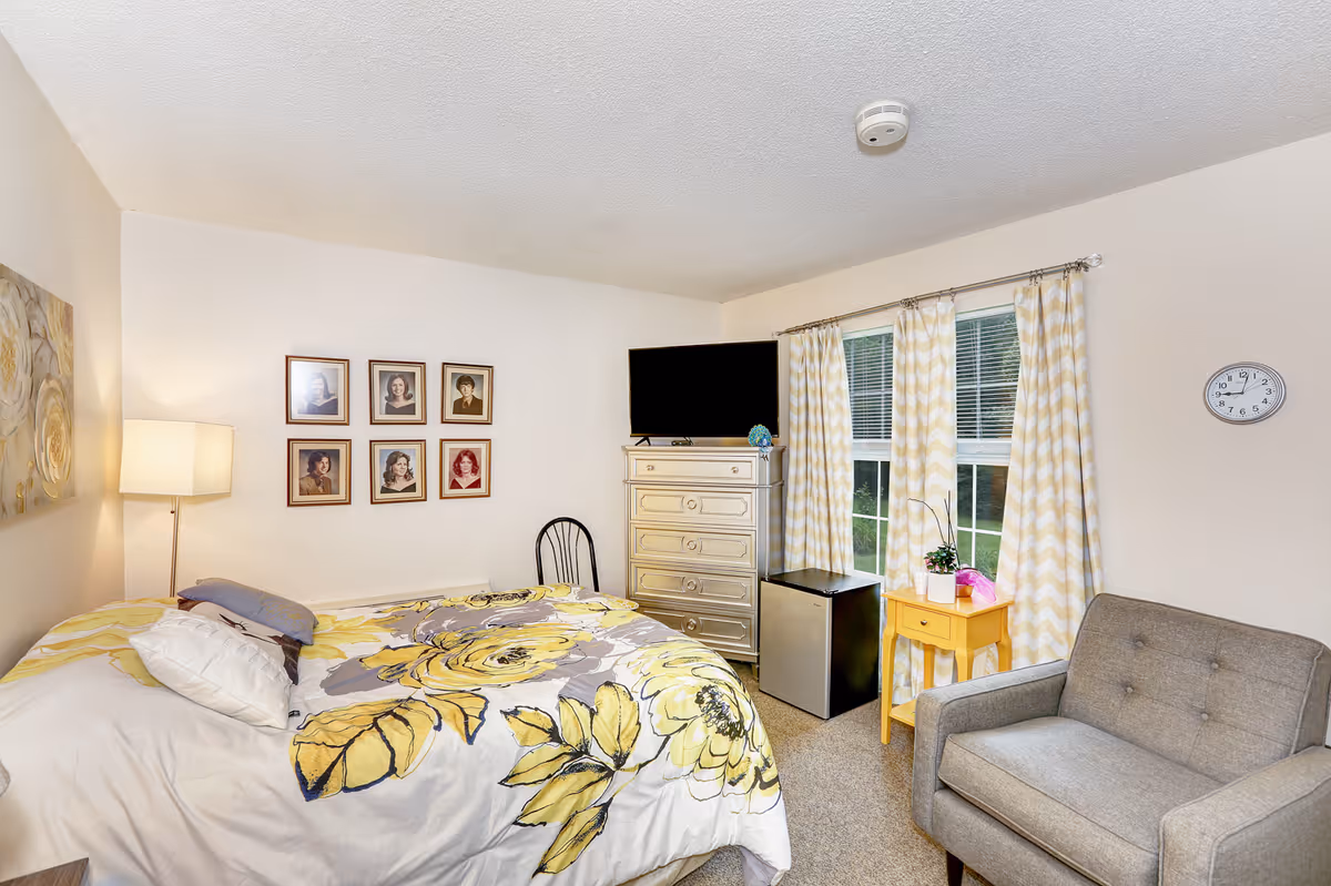 A cozy bedroom with a bed covered in a floral yellow and gray comforter, a gray armchair, a small yellow side table with plants, a dresser with a TV on top, and a window with yellow and white chevron curtains. Six framed portraits hang on the wall above the bed, and a wall clock is visible near the window.