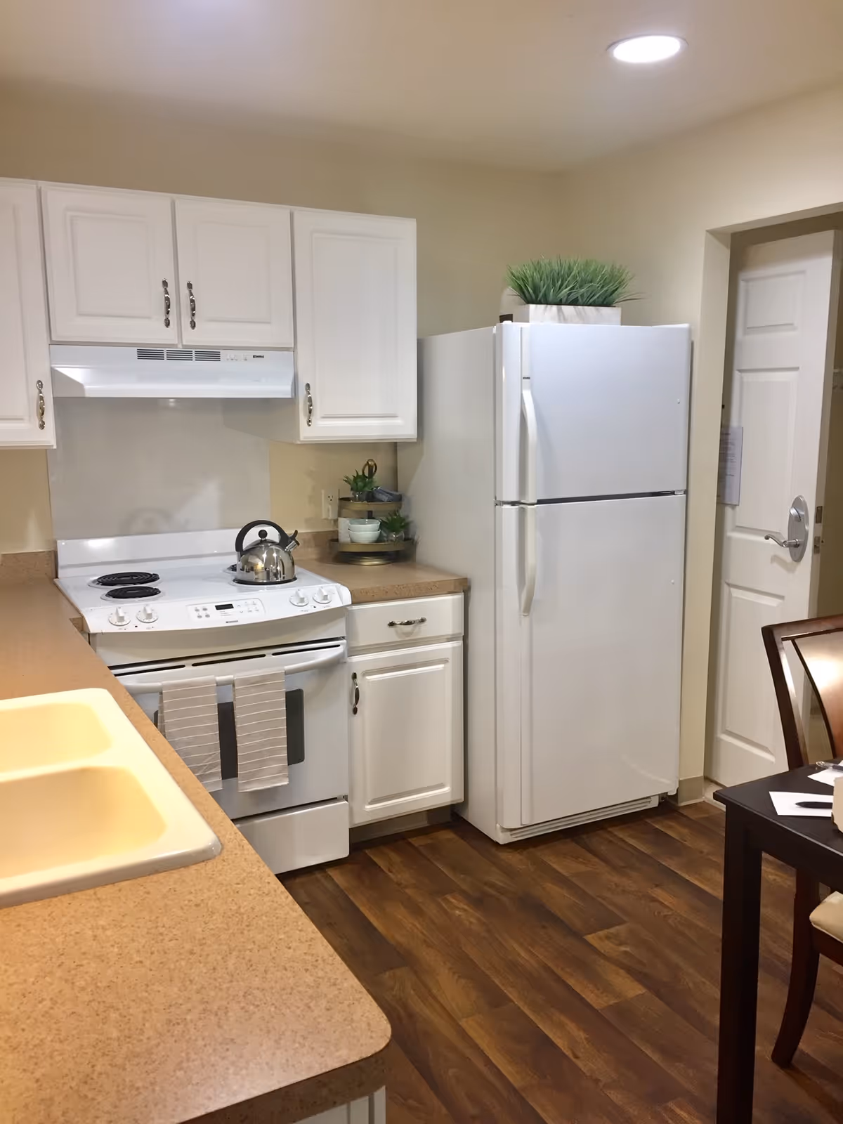 A kitchen with white cabinets, a white electric stove with a kettle on it, a white refrigerator with a potted plant on top, beige countertops, a double sink, and wooden flooring. A dining table with chairs is partially visible on the right side.