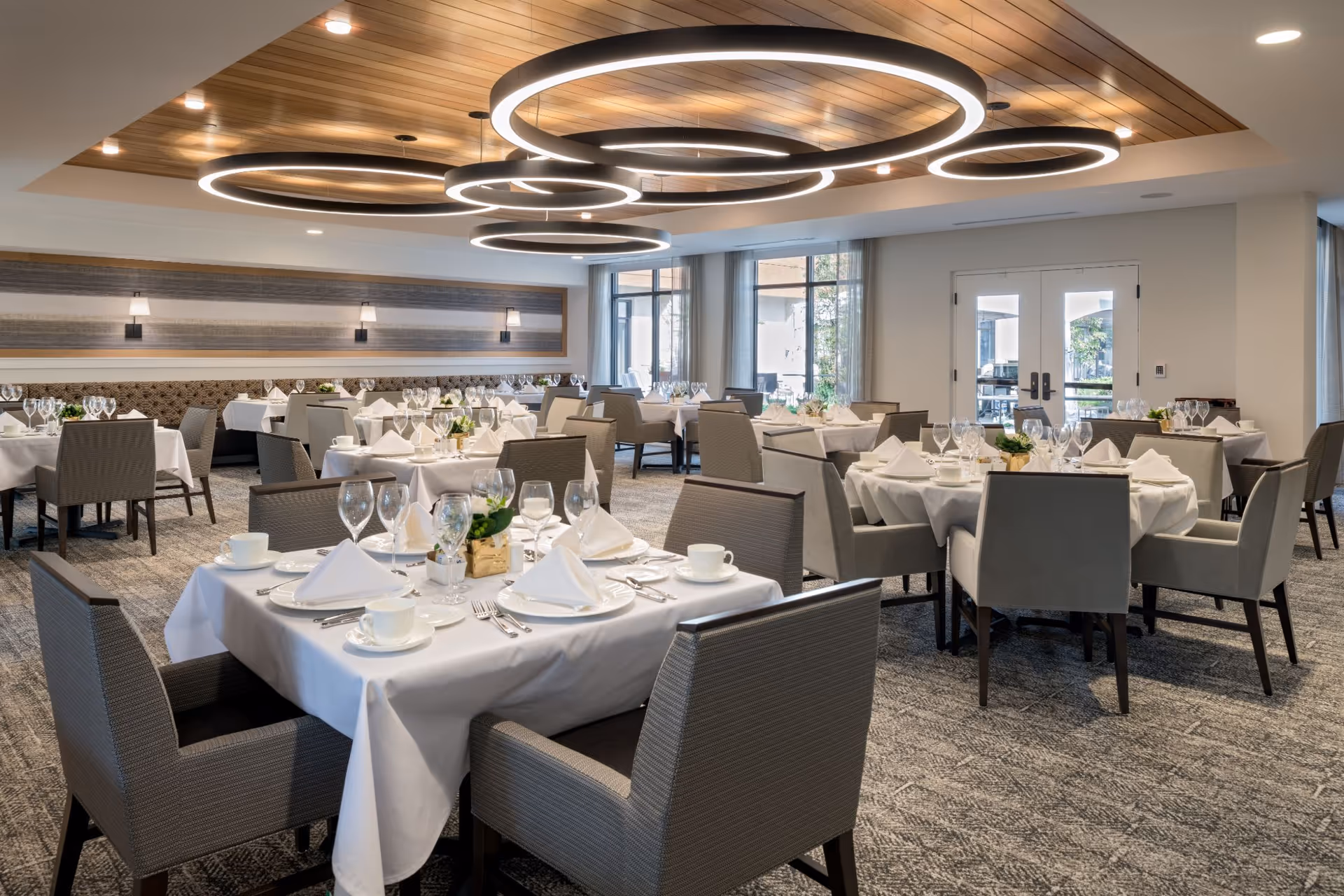 A spacious and elegant dining room with multiple tables covered in white tablecloths, each set with white napkins, plates, cups, and glassware. The room features modern circular ceiling lights, large windows allowing natural light, and comfortable gray upholstered chairs around the tables.