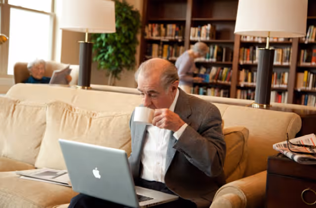 An elderly man sitting on a beige couch in a library or common area, drinking from a white cup while using a silver Apple laptop. In the background, there are bookshelves filled with books and two elderly women, one sitting and reading and the other standing and browsing books. The room is warmly lit with table lamps and has a cozy atmosphere.