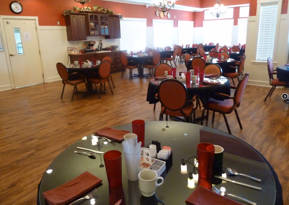 A spacious dining room with multiple round tables set with red napkins, red drinking glasses, white coffee cups, and silverware. The room has wooden flooring, red walls with white trim, large windows with blinds, and chandeliers hanging from the ceiling. There is a cabinet with decorative items and a clock on the wall near a door labeled 'Staff Only Please'.