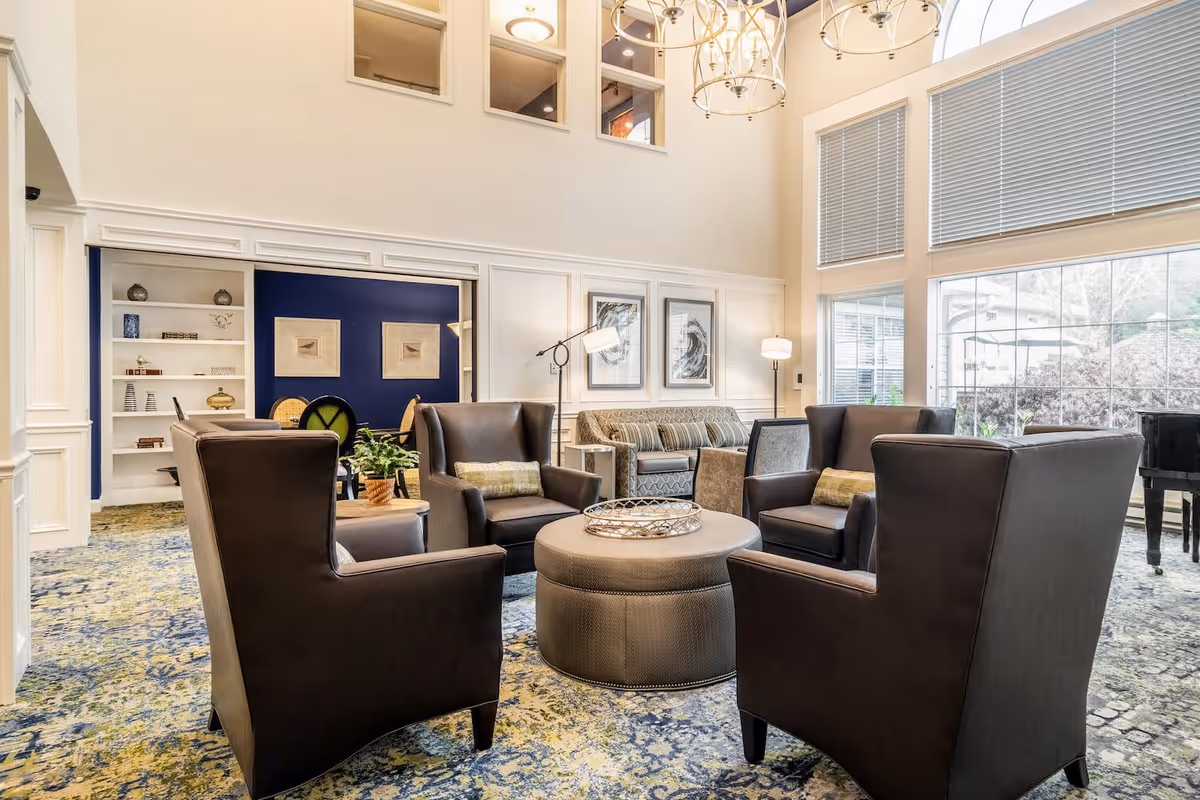 A bright and spacious living room area with four dark brown armchairs arranged around a round ottoman coffee table. Behind the armchairs is a striped sofa and two framed artworks on the wall. Large windows with blinds let in natural light, and a chandelier hangs from the high ceiling. The room features a patterned carpet and a blue accent wall with built-in shelves in the background.