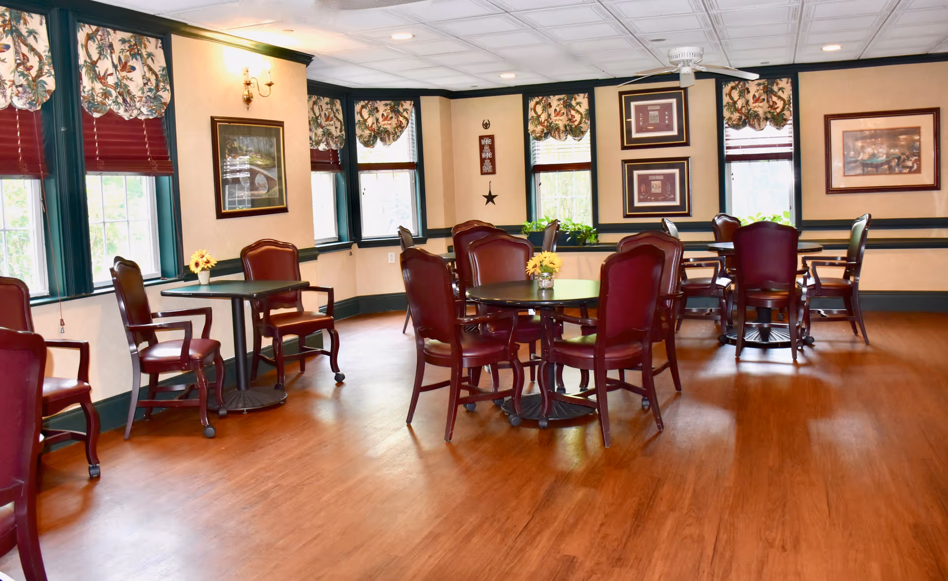 A bright dining room with wooden floors, several round tables surrounded by red cushioned chairs, floral valance curtains on windows, framed artwork on cream-colored walls, and small flower arrangements on the tables.