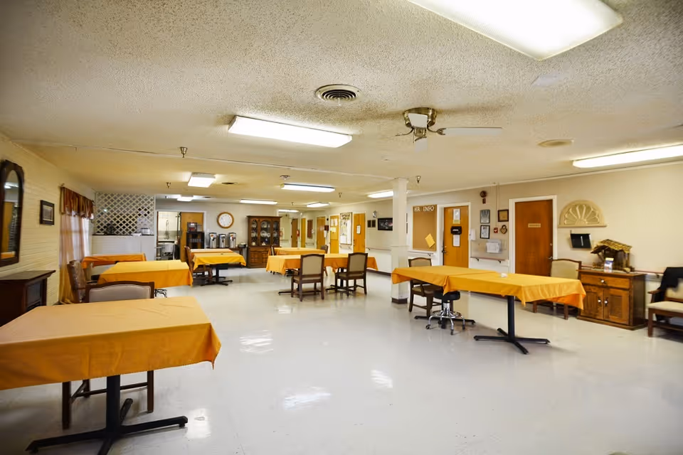 A spacious dining room in a senior living facility with multiple tables covered in orange tablecloths and chairs arranged around them. The room has a white tiled floor, beige walls, fluorescent ceiling lights, and a ceiling fan. There are cabinets, a clock, and a coffee station visible in the background along with doors and bulletin boards on the walls.