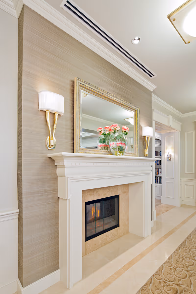 Elegant interior with a white fireplace mantel, mirror, wall sconces, and a vase of pink flowers.