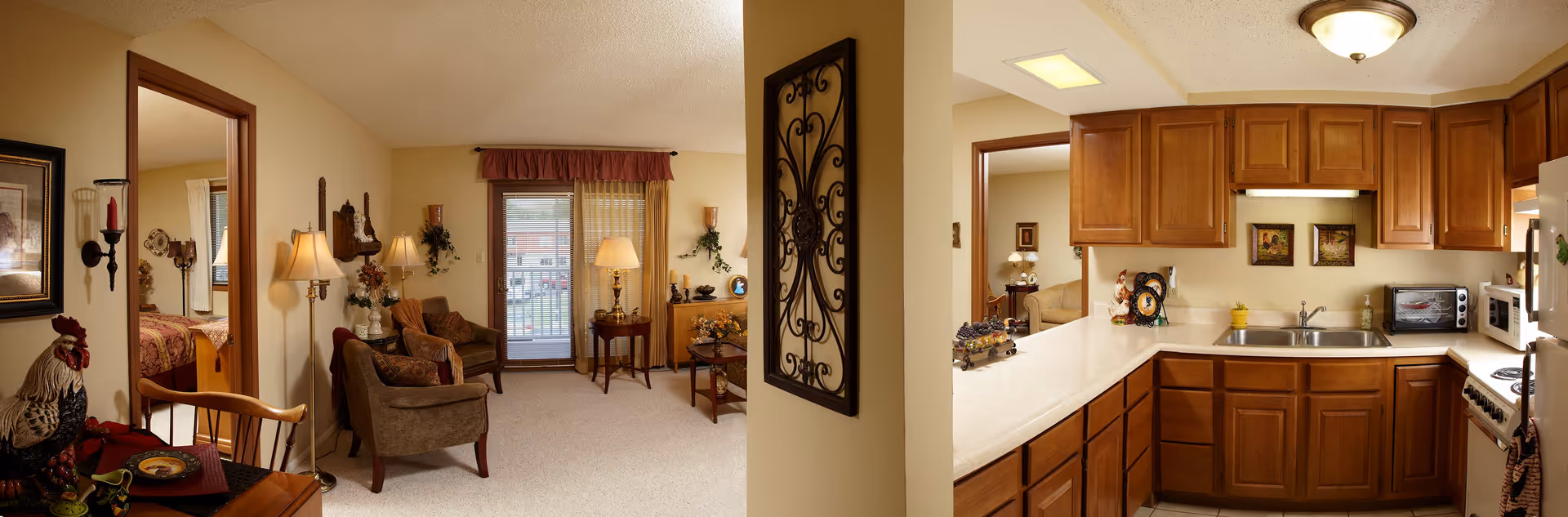 Panoramic interior view of a retirement apartment showing a living room with seating and a balcony door on the left and a kitchen with wooden cabinets and countertops on the right.
