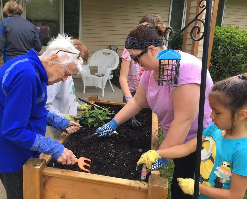 An elderly woman, a middle-aged woman, and two children are gardening together at a raised wooden garden bed outside a building. They are using small gardening tools to work the soil, surrounded by greenery and outdoor furniture.