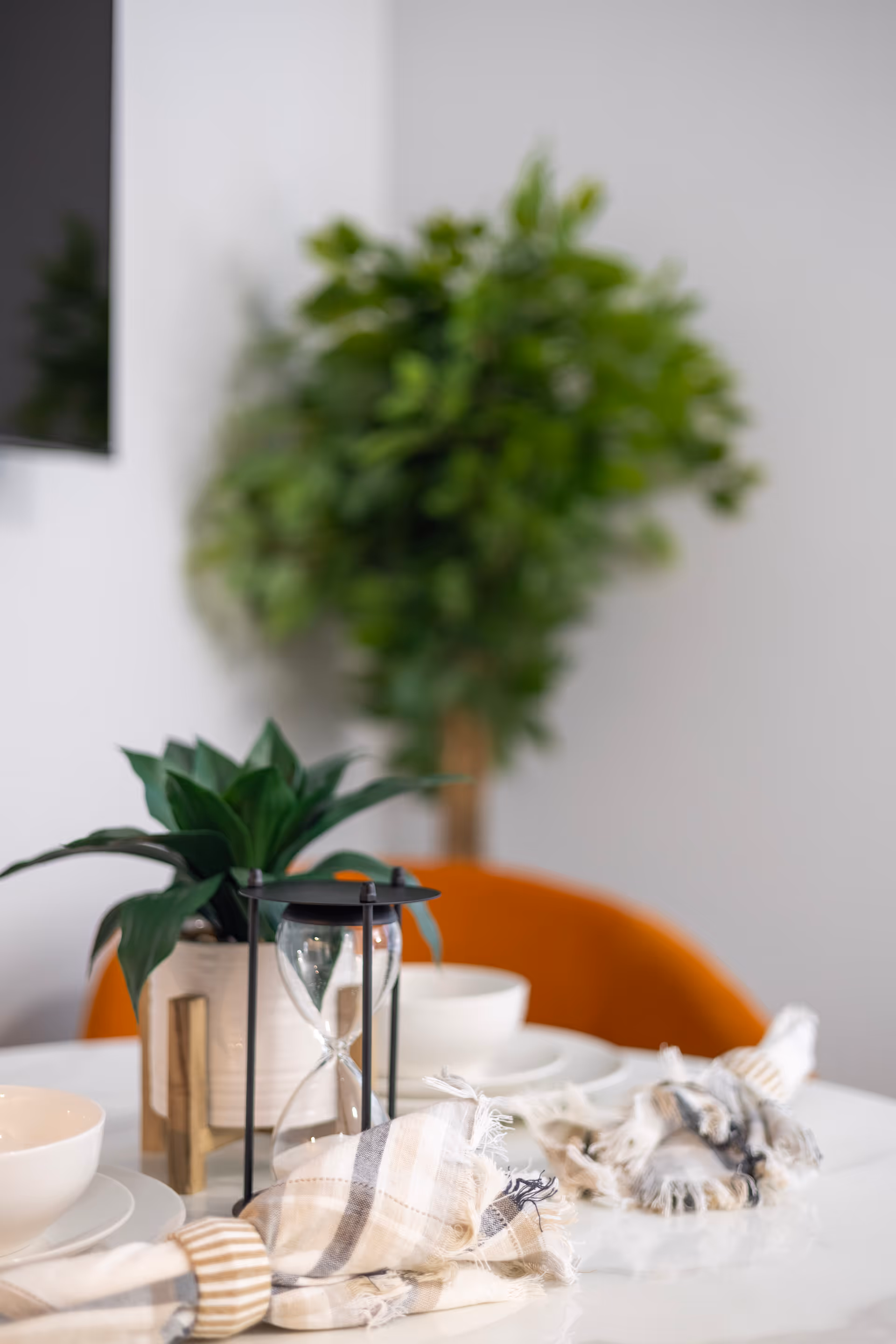 Close-up of a white dining table set with white bowls, striped cloth napkins with napkin rings, a small potted plant, and an hourglass timer. In the background, there is an orange chair and a blurred green leafy plant against a white wall.