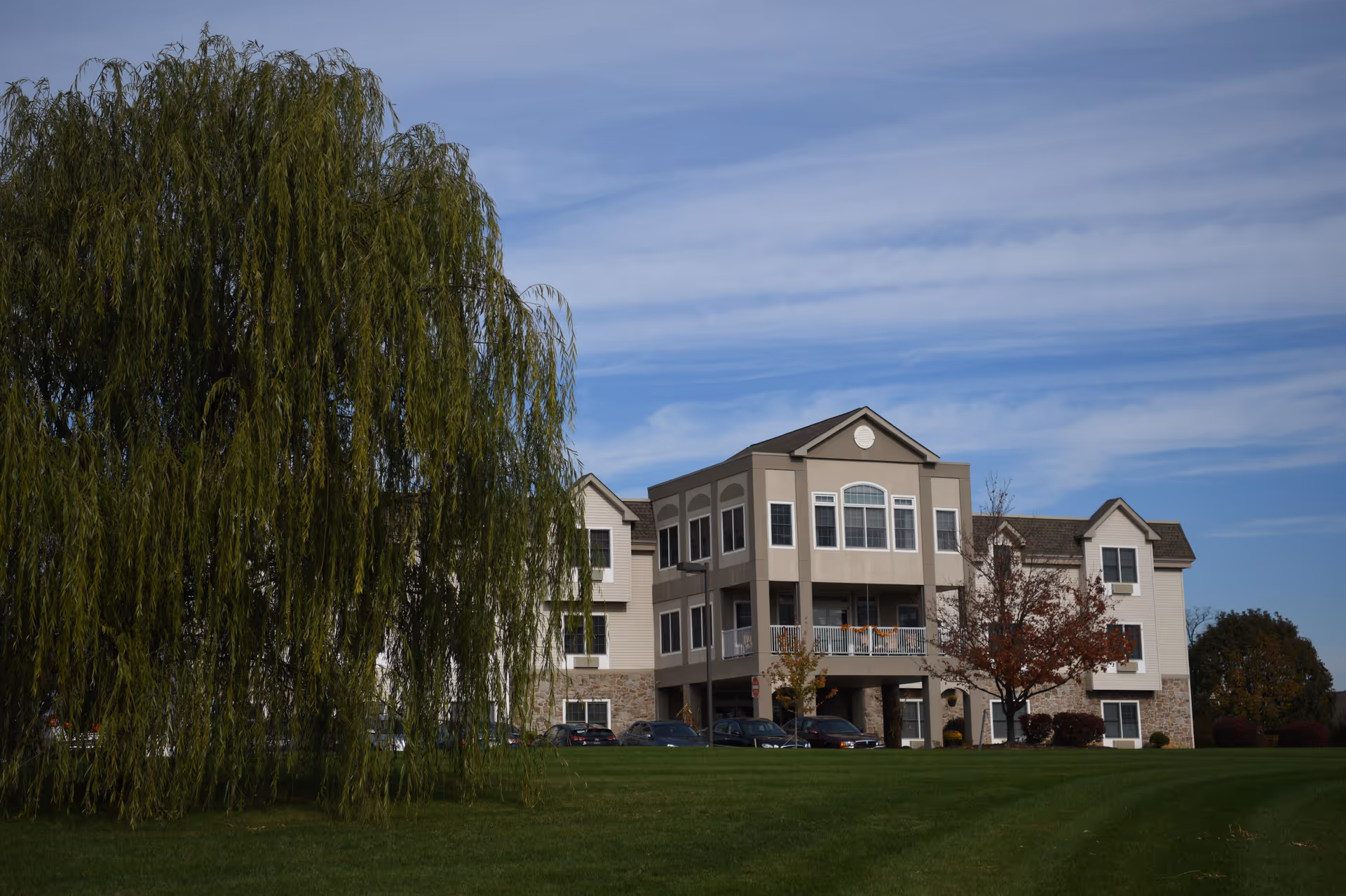 A multi-story senior living facility building with beige and white exterior, large windows, and a balcony. The building is surrounded by green grass, a large weeping willow tree on the left, and a smaller tree with autumn leaves on the right under a partly cloudy blue sky.