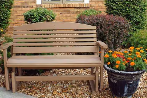 A brown wooden bench sits outdoors beside a brick wall with potted flowers and shrubs.
