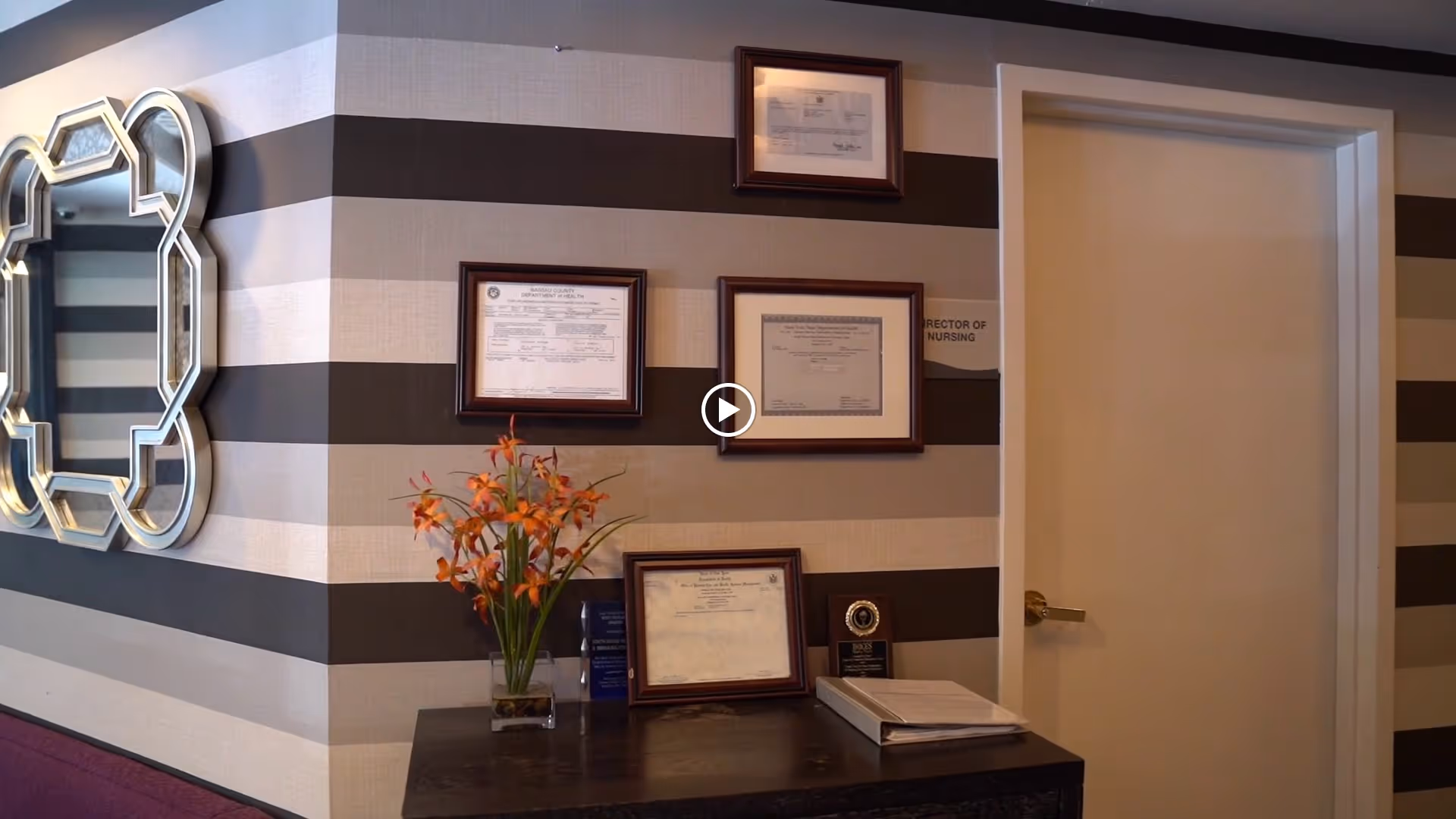 Striped interior wall with framed certificates, a decorative mirror, a vase of orange flowers on a console table and a closed door.