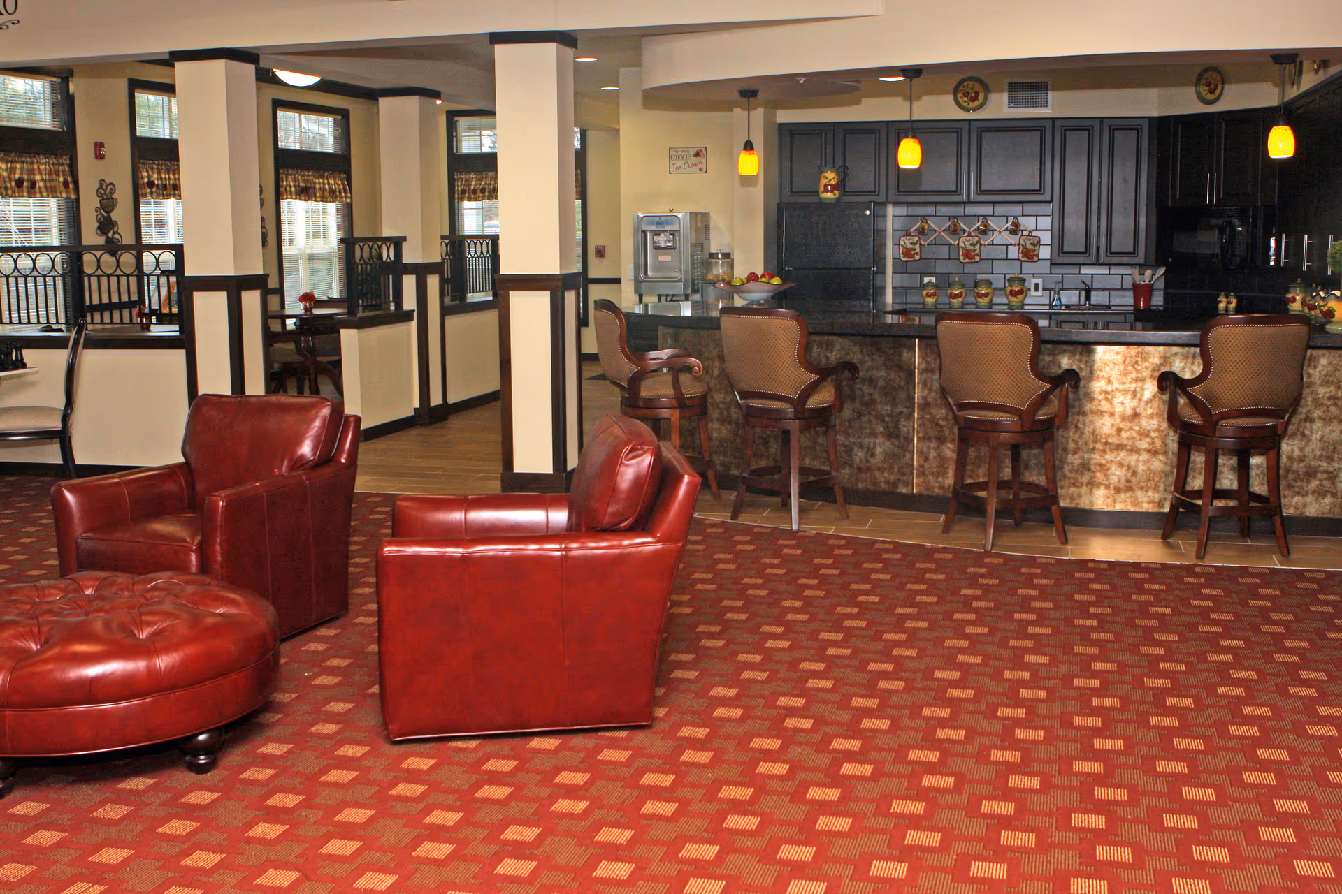 A cozy interior space featuring a seating area with two red leather armchairs and a matching round ottoman on a patterned red carpet. In the background, there is a kitchen area with a counter and four high-backed bar stools. The kitchen has dark cabinetry, a refrigerator, and pendant lights hanging above the counter. The room has several windows with blinds and decorative elements on the walls.