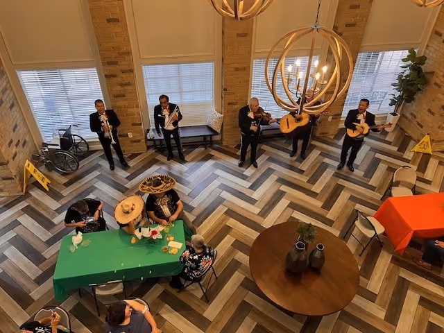 Overhead view of a senior living facility common area with a group of five musicians playing instruments along a brick wall with large windows. Several seniors are seated at tables covered with green and orange tablecloths, some wearing festive hats. The floor has a herringbone pattern, and there are two large circular chandeliers hanging from the ceiling.